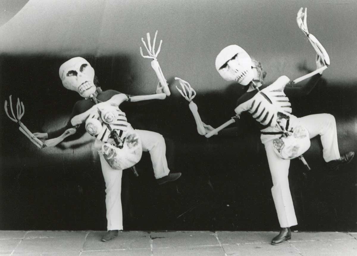 Bare Bones Handspan Theatre black and white picture of 2 skeleton body puppets dancing against an outdoor stone wall