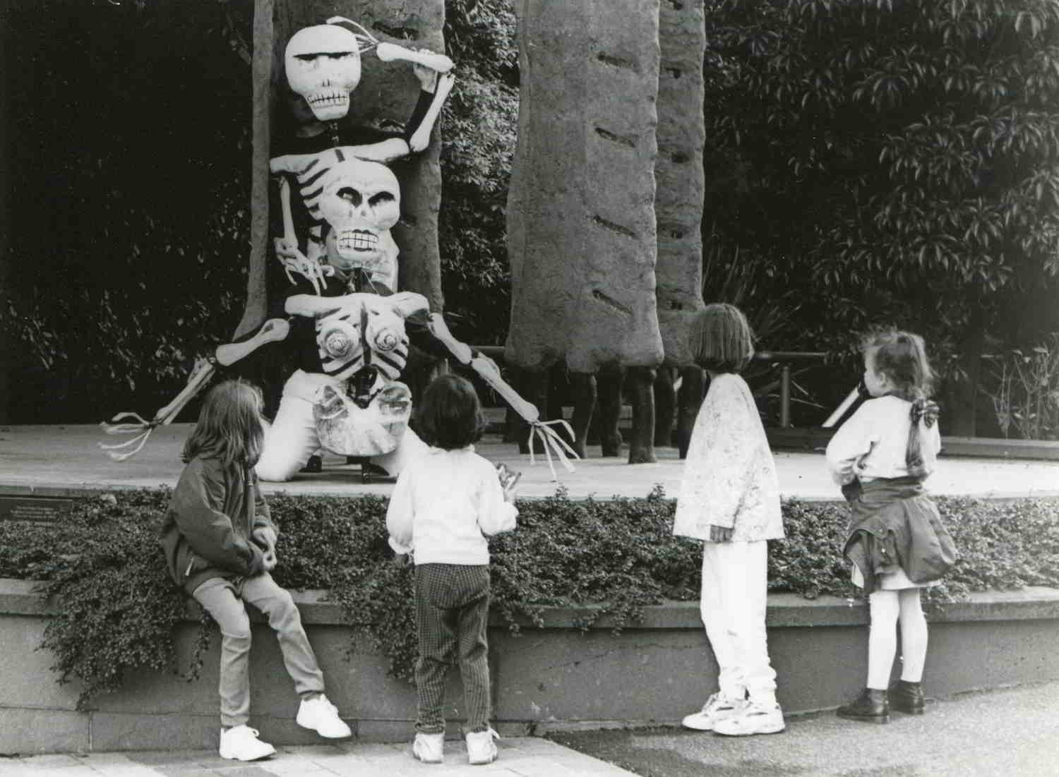 Bare Bones Handspan Theatre 4 children looking at 2 large skeleton puppets leaning at the base of a stone plinth