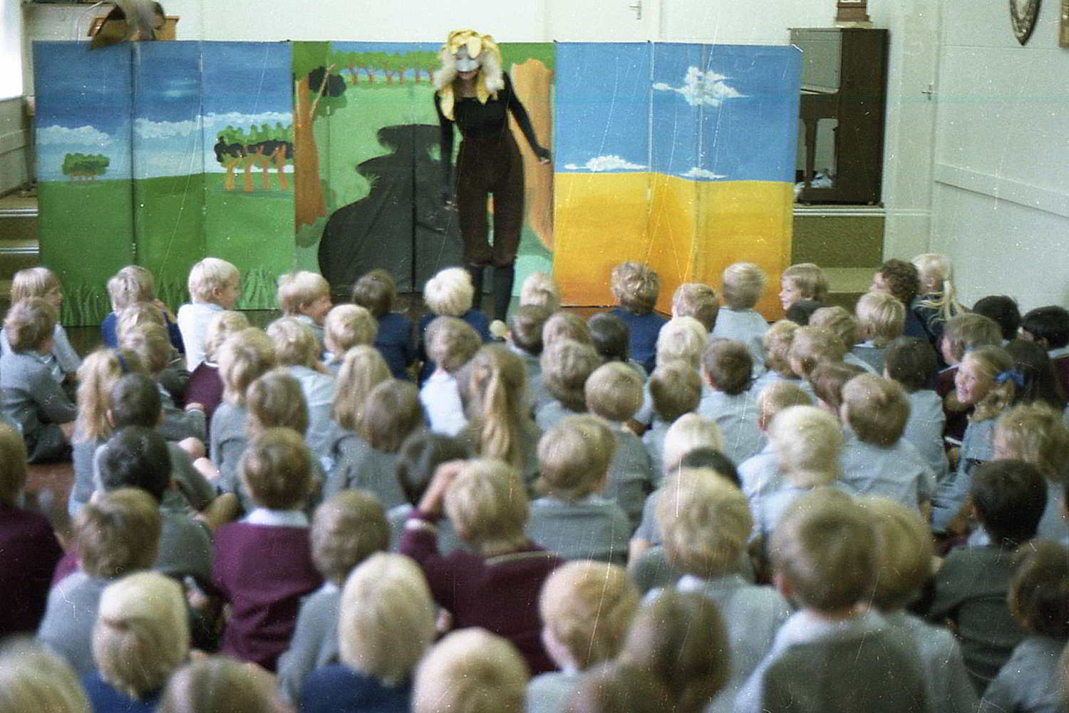 The Bunyip of Berkeley's Creek, Handspan Theatre Actor masked actor in front of stage flats painted with simple landscape, speaking to large group of children in classroom