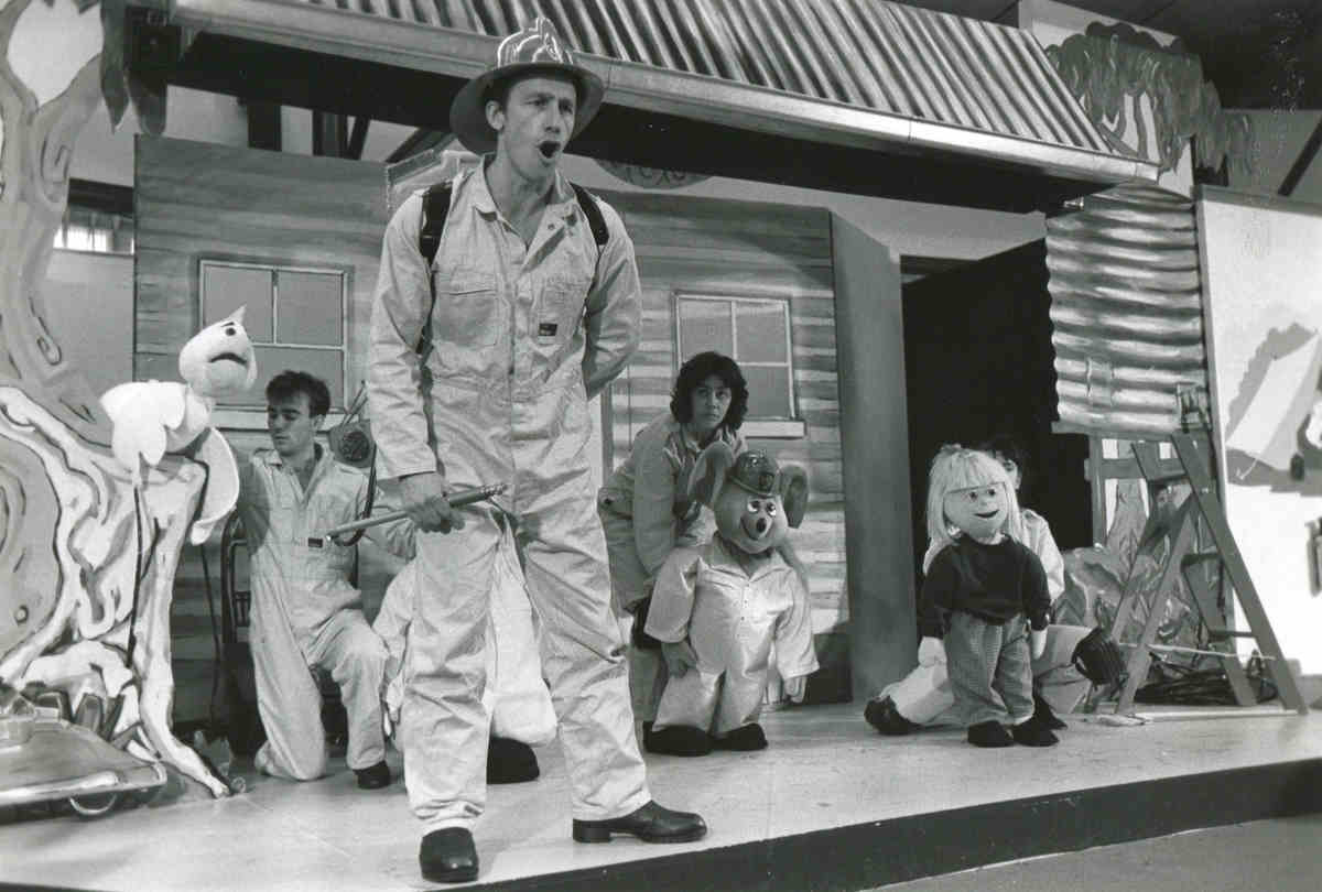Handspan Theatre Captain Koala 3 people onstage with puppets and man in fire-fighter costume standing stage front, singing