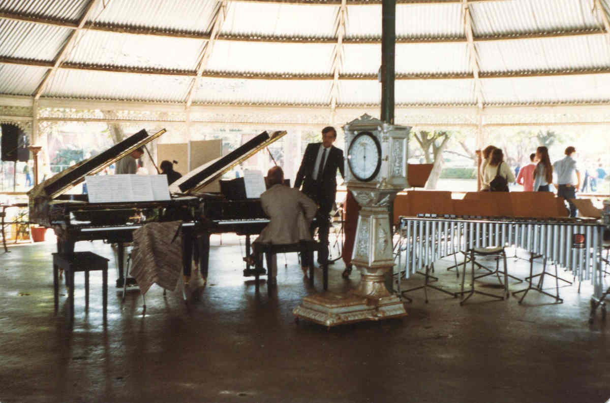 musicians inside park rotunda with 2 pianos and percussion section, old elaborate weighing machine in forground