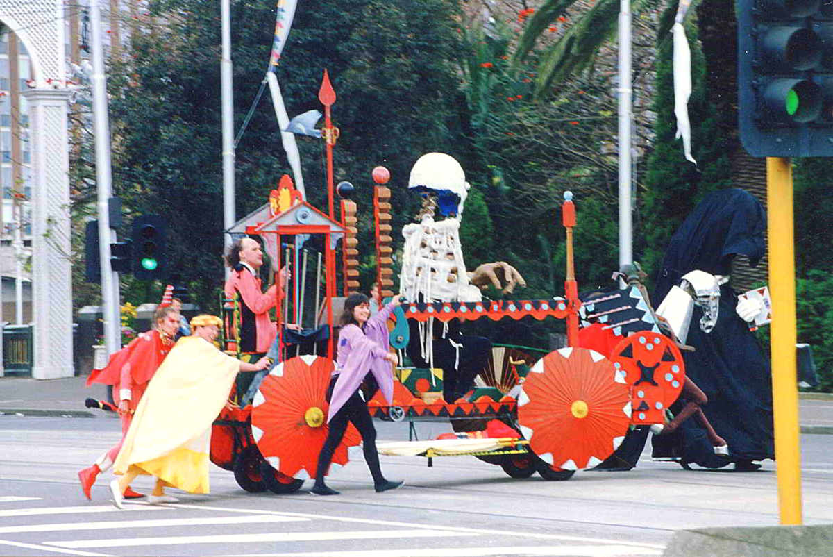 colourful float with red parasol wheels pushed by costumed performers actoss city intersection