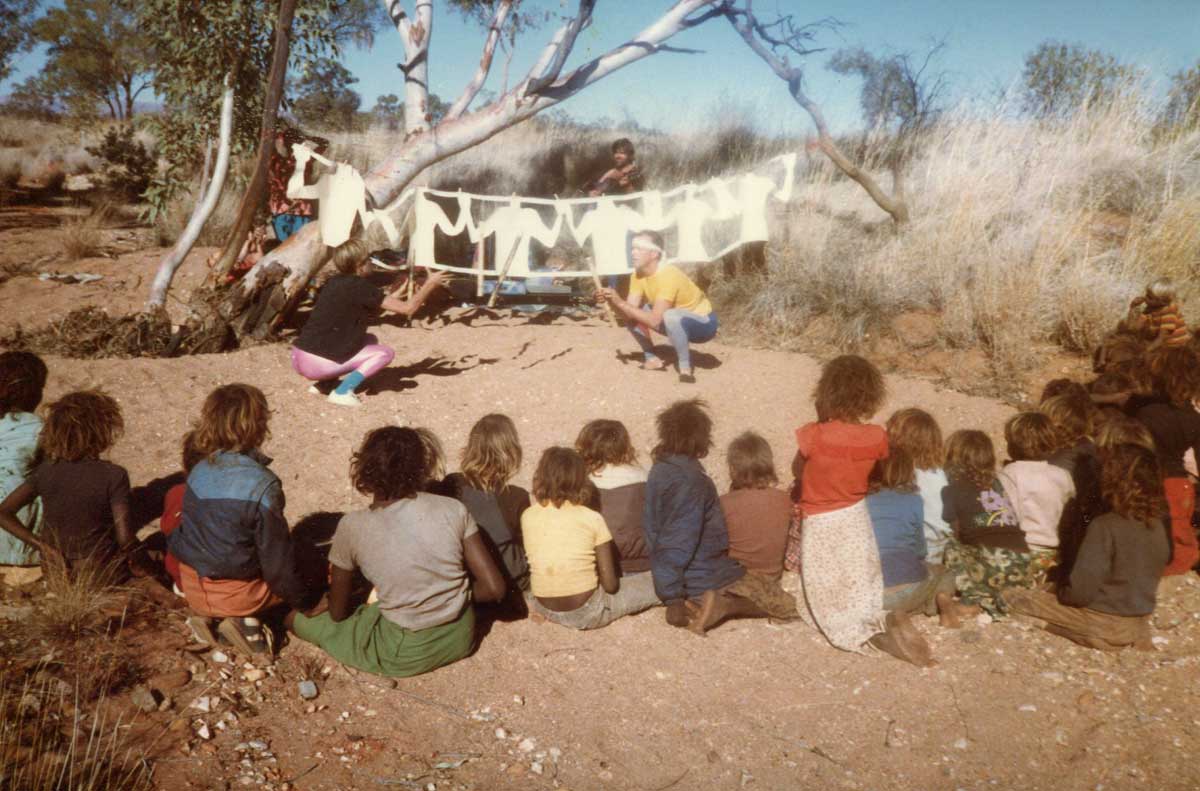 Handspan Theatre Docker River 1985 performers squatting in sandy riverbed with backgrop of cut out white figures and audience of Aboriginal children from behind