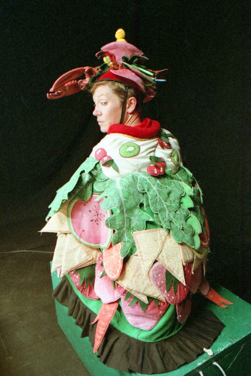 Handspan Theatre Guts costumed actors squatting in embroidered cloak of green leaves and pink vegetables with a lobster helmet