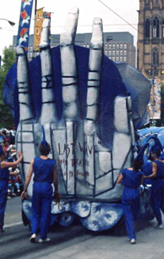 large white hand with words the last wave being pushed along the street by people in blue overalls