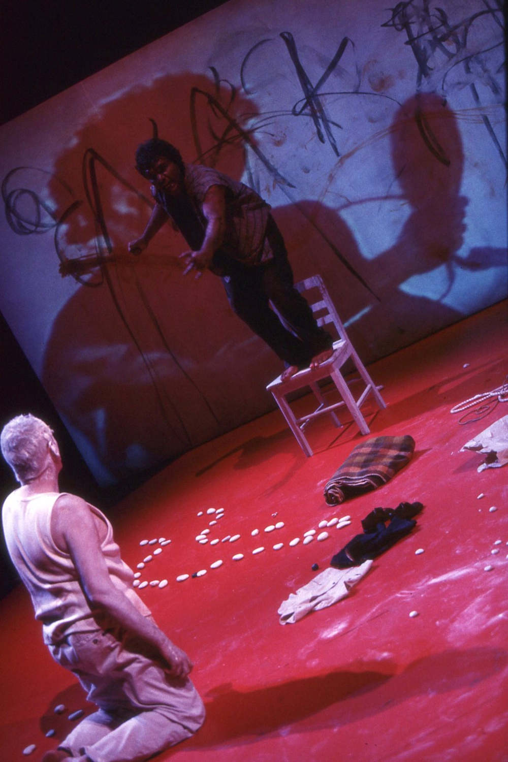 Lift Em Up Socks Handspan Theatre Aboriginal actor standing on chair upstage speaking to a white actor kneeling on red floorcloth beside a white stone pattern
