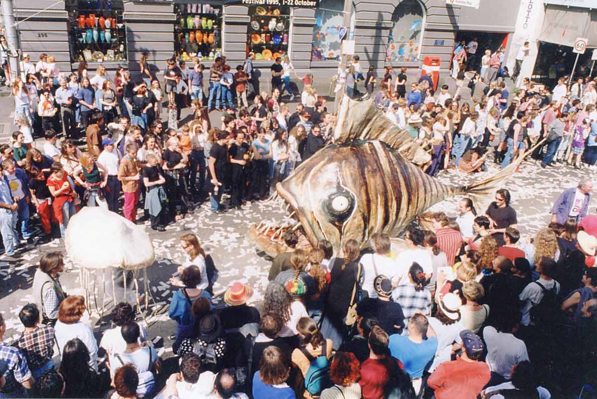 Swim Handspan Theatre large cane fish painted in brown tones following white jelly fish on parade down a crowded city street