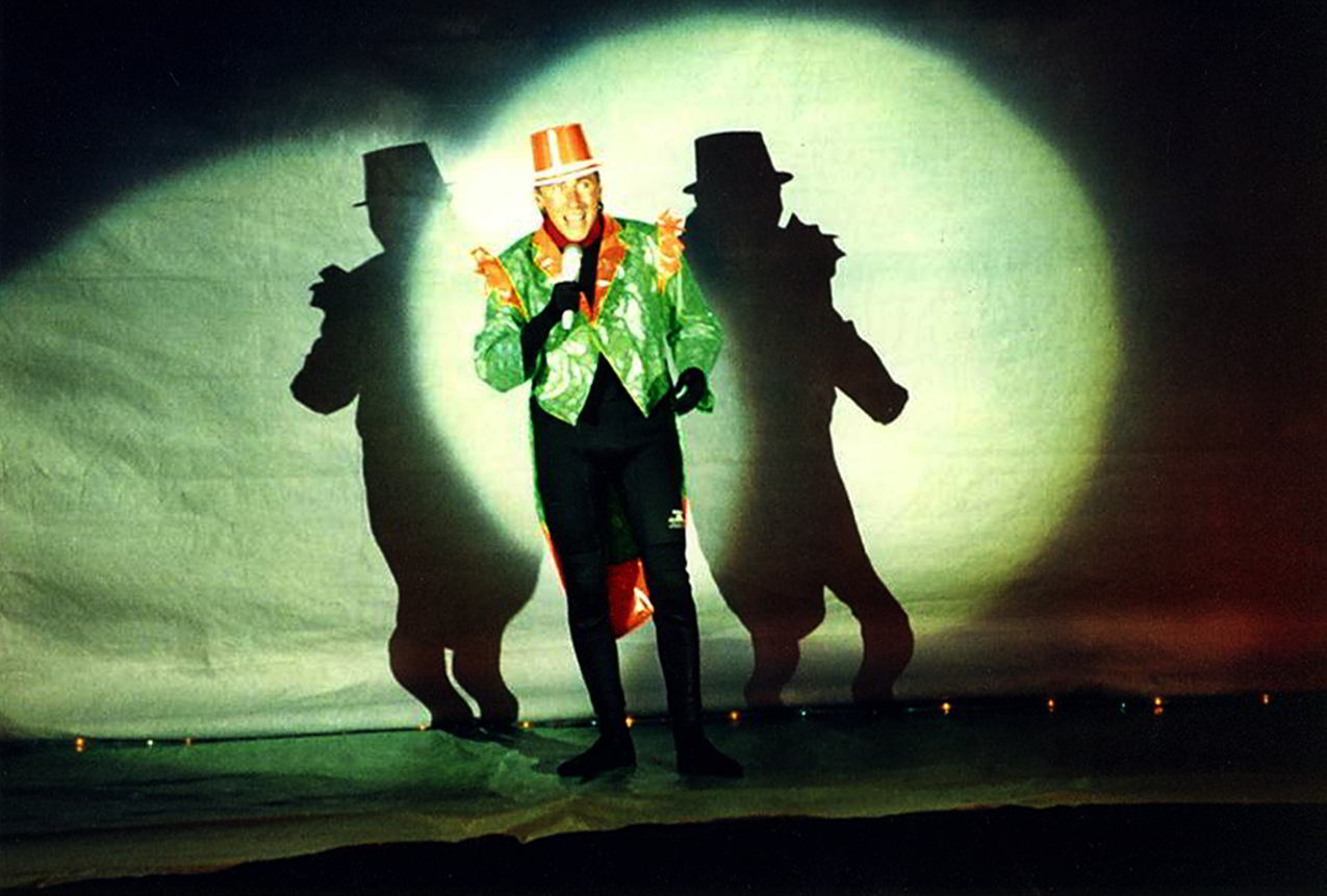 Handspan Theatre Waves of Change Castlemaine Festival figure in orange top hat and green tails in spotlight with double silhouette shadow