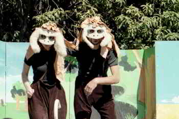 two actors in furry, feathery masks in front of stage flats painted with simple landscape scene