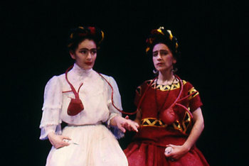 two women sitting side by side, one in red, one in white, each dressed in the style of Kahlo's mexican and german forebears