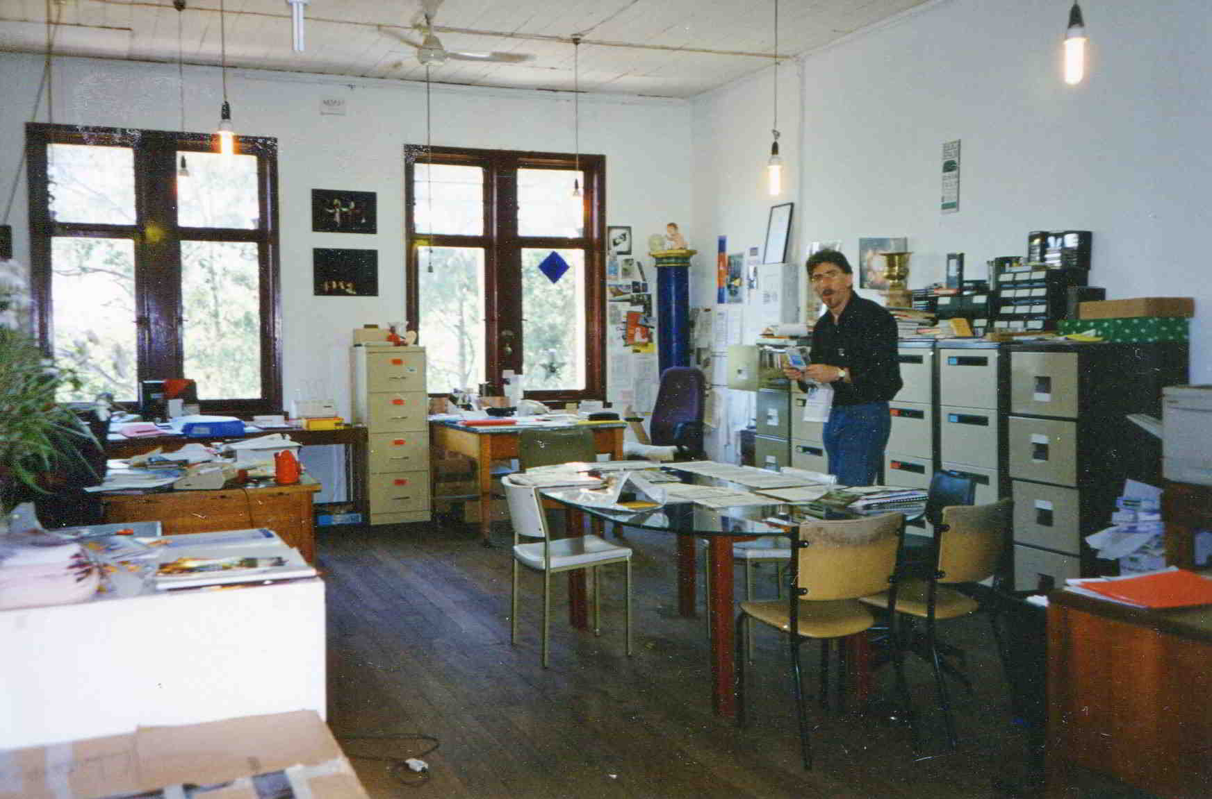 Handspan At Home cluttered office space with large windows, desk corner and central glass table where man is standing
