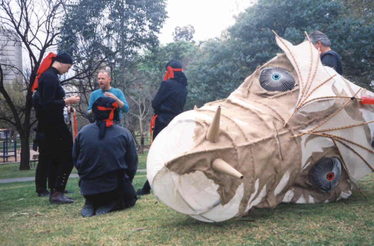 Held on the Breath of the Wind Handspan Theatre 5 people in discussion in a small city park with large white paper and cane dragon head lying on the grass beside them