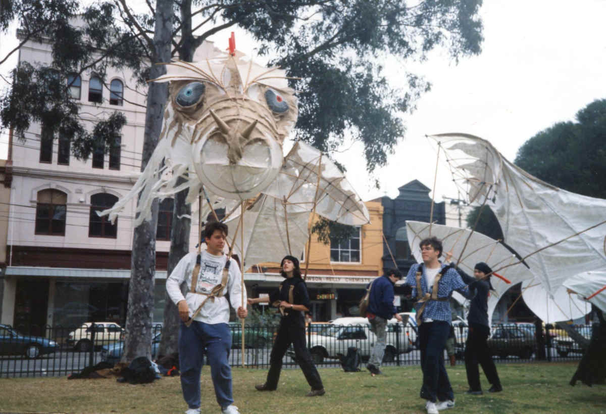 Held on the Breath of the Wind Handspan Theatre 1 person in small city park with large dragon head mounted on body harness, several others with paper and cane dragon wings and tail