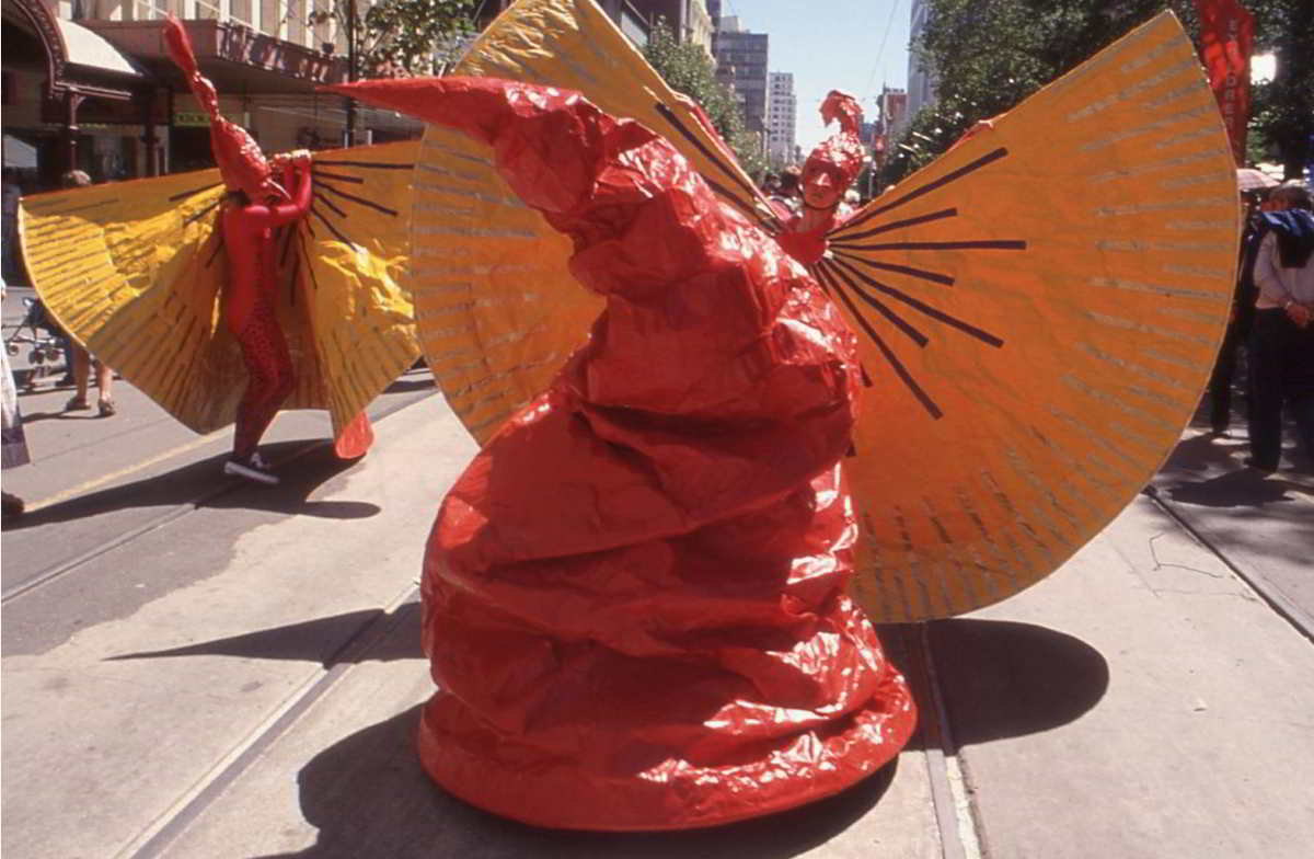 large squashy red cone with red masked figures in large yellow capes in the city street