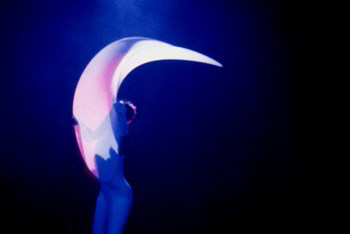 in deep blue light, woman in white body-stocking twists large flexible disc into curved cone shape
